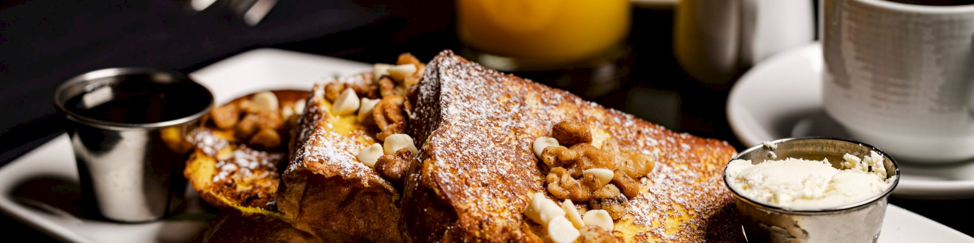 The image shows a delicious plate of French toast topped with nuts, served with a side of cream, syrup, orange juice, and a cup of coffee.